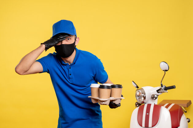 前面正面图：戴着黑色面具的男信使拿着咖啡 戴着黄色的工作帽——大流行服务递送工作修理工帽子送货