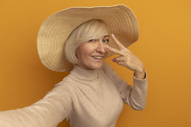 海滩微笑的金发斯拉夫女人 戴着沙滩帽 打着胜利手势 看着橙色的相机时尚姿势手