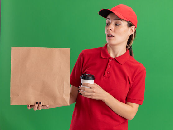 包装一个穿着制服的年轻漂亮的送货女孩拿着纸杯 看着绿色的纸包装制服人年轻