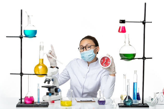 实验室前视图：身穿医疗服的女化学家 戴着面罩 手持浅白色背景上的红色时钟病毒实验室化学冠状病毒-飞溅化学前面西装