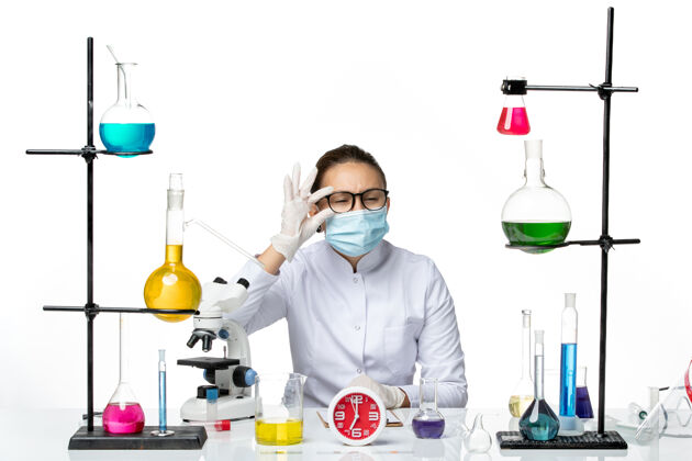 医生前视图穿着医疗服的女化学家戴着面罩坐着 拿着溶液看着白色背景的化学家实验室病毒-飞溅药物病毒化学家