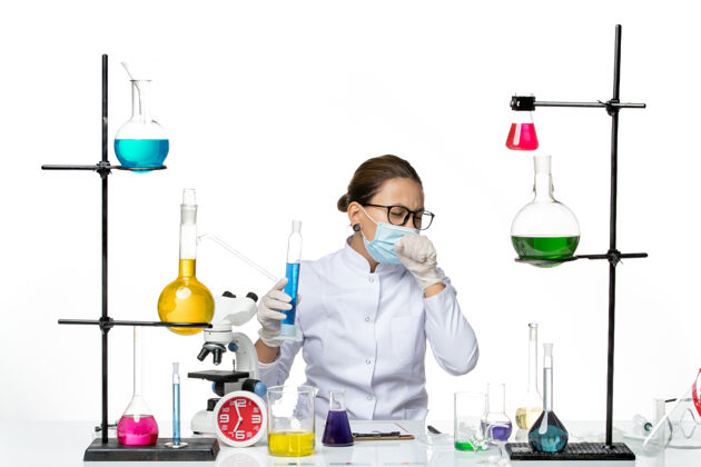 正面前视图穿着医疗服的女化学家带着面罩拿着浅白色背景上蓝色溶液的烧瓶溅起病毒化学实验室的病毒医生病毒容器