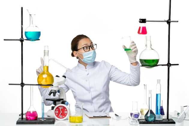 化学前视图穿着医疗服的女化学家 带着面罩 在浅白的背景上拿着绿色溶液 飞溅着实验室病毒化学的covid-溶液喷溅外套