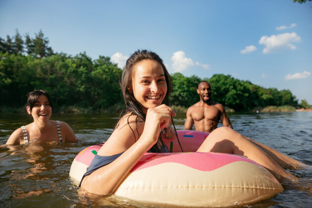 新鲜一群快乐的朋友在河里欢笑和游泳在阳光明媚的日子里 快乐的男人和女人拿着橡胶圈在河边做甜甜圈夏日 友谊 度假 周末的概念玩冒险团体