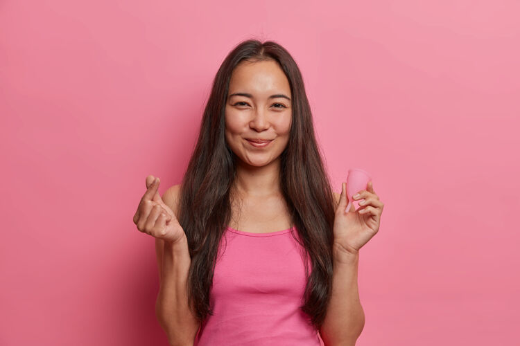 内年轻的亚洲黑发女人拿着月经杯摆姿势 展示如何使用 给出建议 做出韩式的招牌 隔离在粉色的墙上女人健康 零废物替代和妇科清洁药品穿