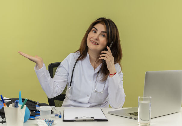 穿微笑的中年女医生穿着医用长袍和听诊器坐在办公桌旁 拿着医疗工具和笔记本电脑打电话 显示空手孤立空听诊器中年