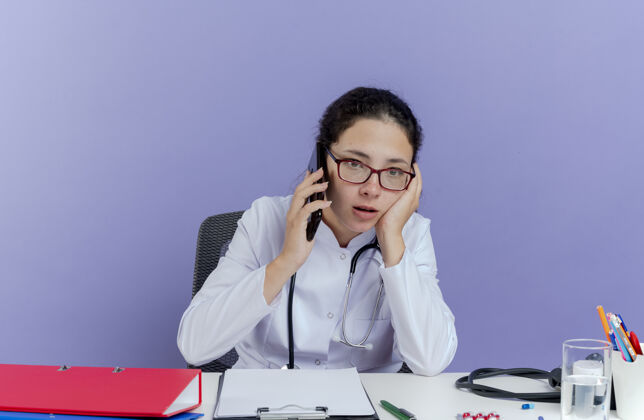 长袍令人印象深刻的年轻女医生穿着医用长袍和听诊器坐在办公桌旁 拿着医疗工具讲着电话 表情直勾勾地抚摸着孤立的脸年轻女士印象