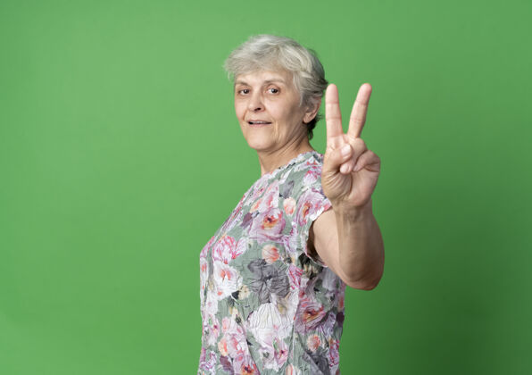 老人高兴的老妇人侧着身子站着 指着绿色墙上孤立的胜利手势手老人绿色