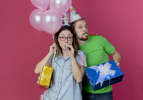 男人快乐的年轻人戴着派对帽拿着礼盒吹着口哨站在惊讶的年轻女孩后面拿着氦气球和礼盒隔离在粉红色的墙上气球帽子粉色