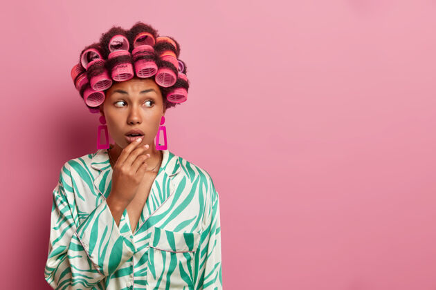 优雅黑皮肤女人的照片卷发 戴卷发器 在家里做发型 手放在张开的嘴上 穿着休闲服 对着粉色的墙壁摆姿势 旁边空无一人罗西肖像戒指
