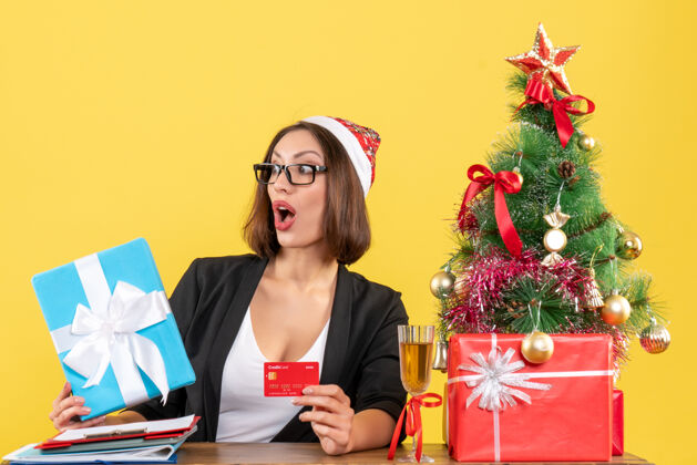 快乐惊喜迷人的女士身着西装 戴着圣诞老人的帽子和眼镜 在黄色的办公室里展示礼物和银行卡惊喜圣诞老人圣诞老人