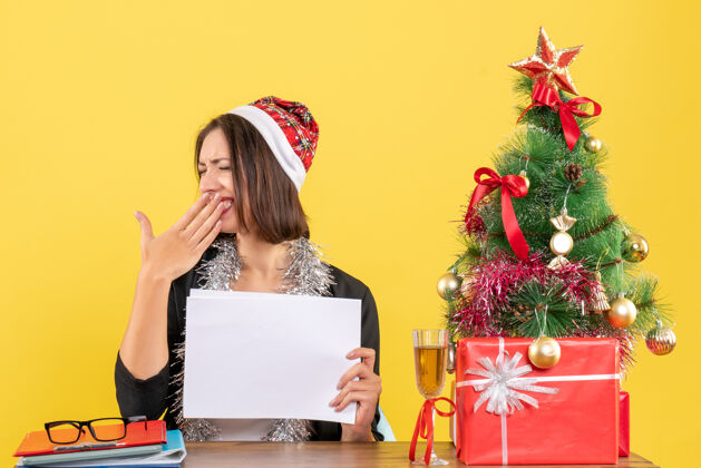 抱着一位身着西装 戴着圣诞老人帽 戴着新年装饰品 面带微笑的商务女士独自工作 手里拿着文件 打着哈欠 坐在一张桌子旁 桌子上放着圣诞树坐着生活工作