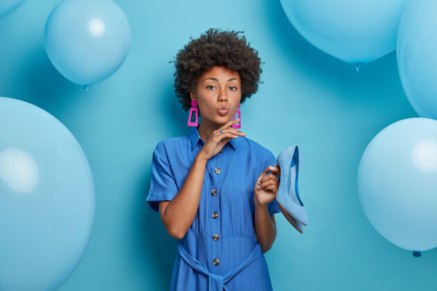 非洲女人 时尚 风格 派对理念优雅严肃的女人穿着蓝色的裙子 拿着高跟鞋 穿着主题派对的衣服 准备出门 拿着氦气球对着蓝色的墙壁摆姿势凝视高跟鞋单色