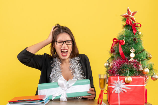 树情绪紧张的商务女士穿着西装 戴着眼镜 手里拿着礼物 坐在一张桌子旁 桌子上放着圣诞树坐人时尚