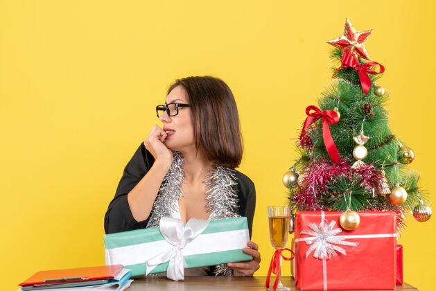 情感情绪化的商务女士穿着西装 戴着眼镜指着她的礼物 坐在一张桌子旁 桌子上放着一棵圣诞树指向快乐头发