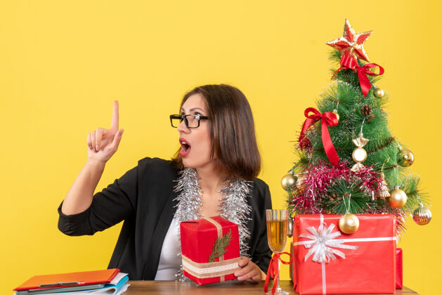 套装好奇的商务女士穿着西装 戴着眼镜 手里拿着礼物 指着桌子坐在办公室里 桌子上放着圣诞树眼镜站起来微笑