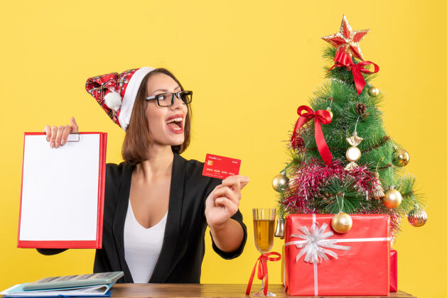 成人集中迷人的女士西装与圣诞老人帽子和眼镜显示银行卡和文件在黄色的办公室隔离漂亮快乐西装