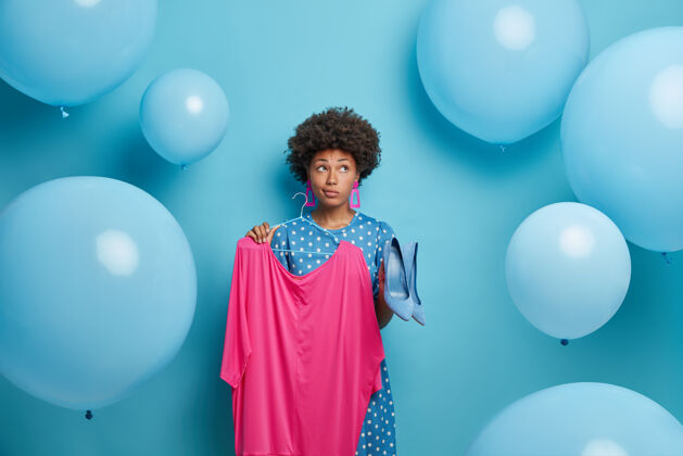 民族体贴的女性聚会者为特殊场合选择喜庆的衣服 衣架上拿着粉色的连衣裙和高跟鞋 表情沉思 孤零零地站在蓝色的墙上 周围放着气球气球表情姿势