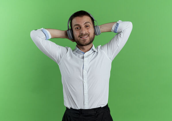 耳机戴着耳机的帅哥高兴地把手放在头后 隔离在绿色的墙上手帅哥拜托