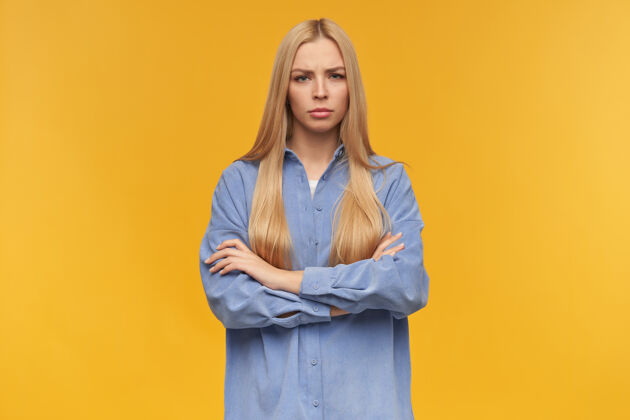 严肃严肃的 金发的成年女孩的肖像穿着蓝色衬衫人和情感的概念双臂交叉在胸前看着相机 橙色背景下孤立青少年面部情绪