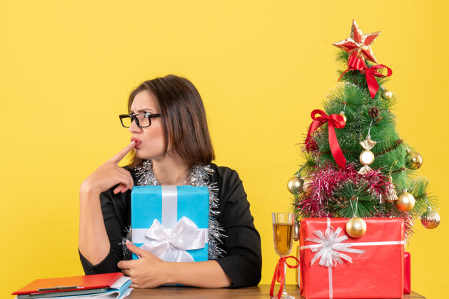 树一位穿着西装 戴着眼镜 拿着礼物 坐在一张桌子旁 桌上放着圣诞树的商务女士大吃一惊人眼镜时尚