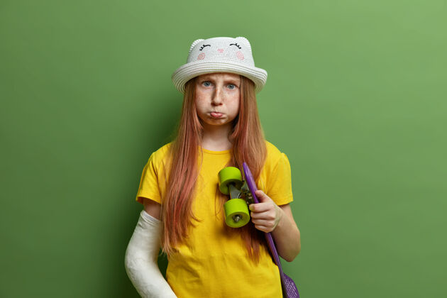 忧郁骑滑板后手臂受伤的红发女孩 骨折 闷闷不乐的表情 长长的红发 穿着夏天的衣服 隔离在绿墙上极限运动 儿童 生活方式悲伤雀斑情绪