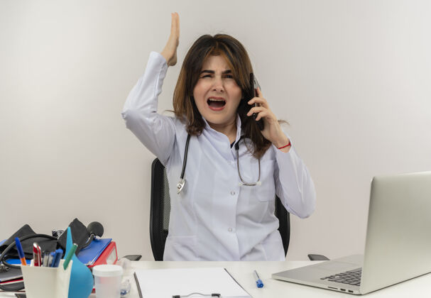 人愤怒的中年女医生穿着医用长袍和听诊器坐在办公桌前拿着医疗工具剪贴板和笔记本电脑讲电话举起手来电话女人工具