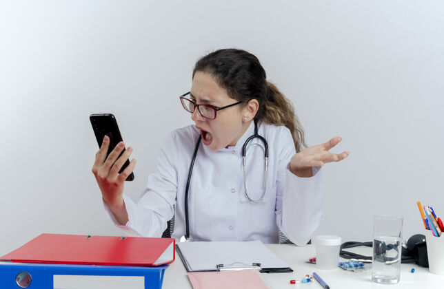 穿愤怒的年轻女医生穿着医用长袍 戴着听诊器和眼镜坐在办公桌旁 手持医疗工具 看着手机 显示空手孤立表演桌子长袍