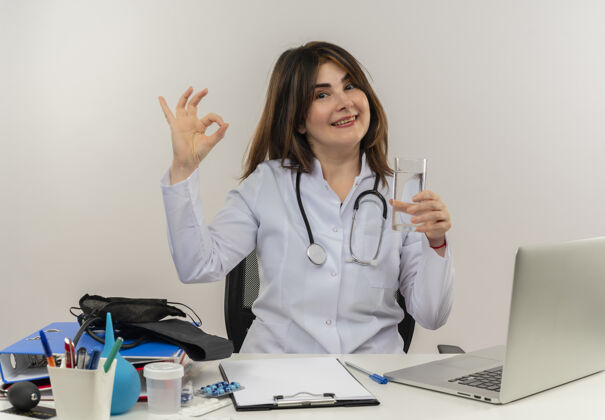 中年微笑的中年女医生穿着医用长袍和听诊器坐在办公桌旁 拿着医疗工具 剪贴板和笔记本电脑 拿着一杯隔离水做着ok的手势坐着长袍女人