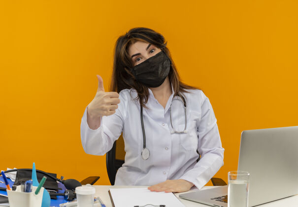 拇指自信的中年女医生戴着医用口罩坐在办公桌旁 带着医疗工具剪贴板和笔记本电脑 手放在办公桌上 大拇指向上伸出女人面具医疗