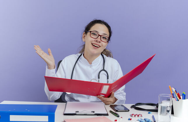 穿快乐的年轻女医生穿着医用长袍和听诊器坐在办公桌旁拿着医疗工具拿着文件夹看着显示空手孤立桌子抱着医生