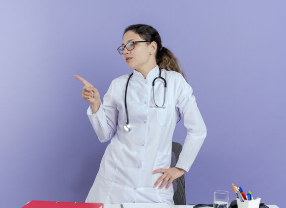 女人年轻的女医生穿着医用长袍 戴着听诊器 戴着眼镜 站在桌子后面 手里拿着医疗工具 手放在腰上 看着一边紫色站穿