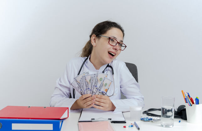 女人印象深刻的年轻女医生穿着医用长袍 戴着听诊器和眼镜 坐在办公桌旁 拿着医疗工具 拿着钱 一边看一边隔离长袍医疗抱着
