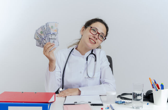 医生面带微笑的年轻女医生穿着医用长袍 戴着听诊器和眼镜 坐在办公桌旁 手里拿着医疗工具 手里拿着钱 孤零零地抬头看人成年人医疗