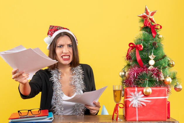 坐着情绪激动 愤怒的商务女士穿着西装 戴着圣诞老人的帽子和新年装饰品 手里拿着文件 坐在桌子旁放着圣诞树愤怒树圣诞老人