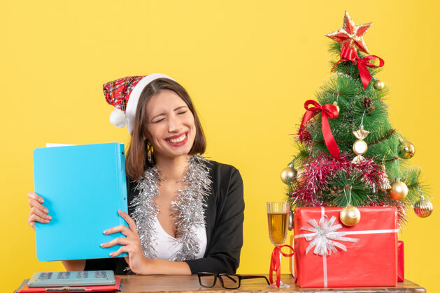 成人心满意足的快乐迷人的女士 穿着西装 戴着圣诞老人的帽子和新年的装饰品 手里拿着文件 在黄色的办公室里与世隔绝文件性感西装