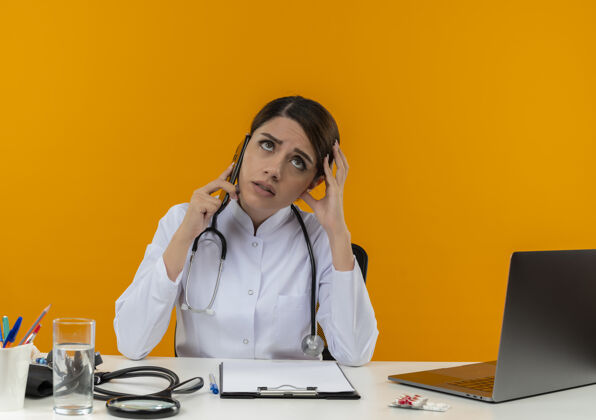 女性年轻的女医生穿着医用长袍和听诊器坐在办公桌旁 拿着医疗工具和笔记本电脑打电话 摸着头孤立地抬头看黄色穿着触摸