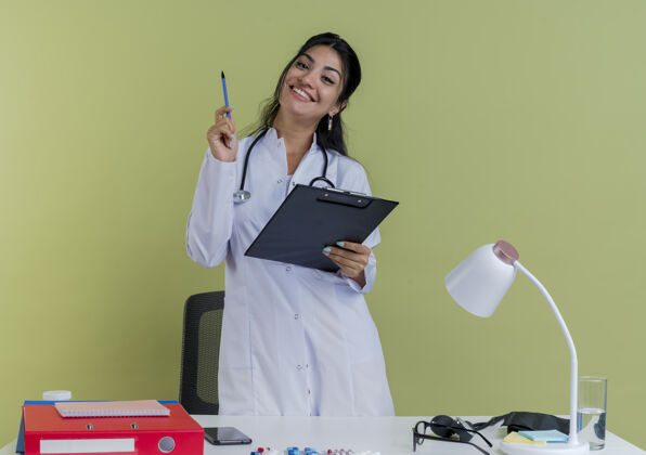 剪贴板微笑着的年轻女医生穿着医用长袍和听诊器站在桌子后面 手里拿着医疗工具 手里拿着剪贴板和笔女士穿着听诊器