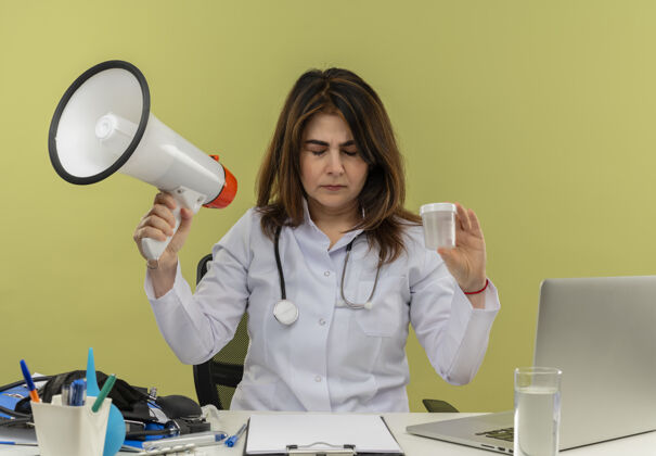人中年女医生穿着医用长袍和听诊器坐在办公桌旁 手持医疗工具和笔记本电脑 手持扬声器和医用烧杯 闭眼隔离桌子绿色工具