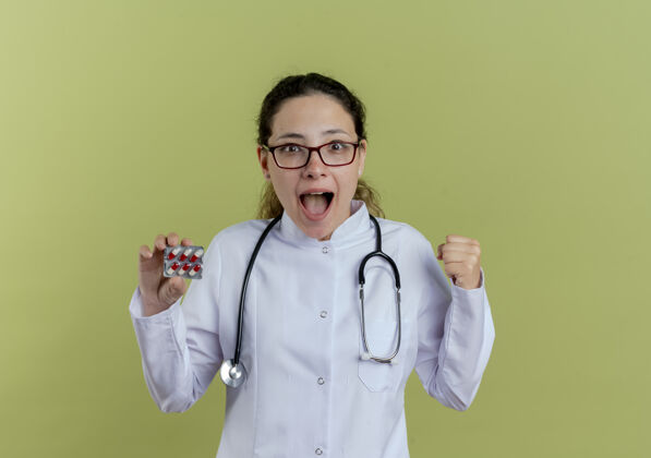 人快乐的年轻女医生 穿着医用长袍 戴着听诊器 戴着眼镜 拿着药片 做着“是”的手势是医生表演