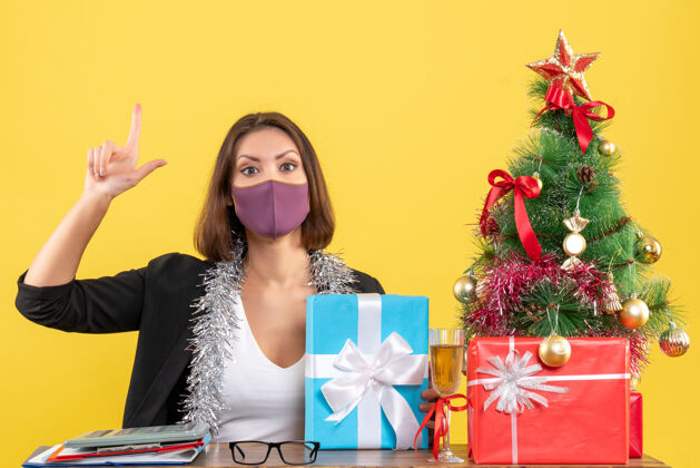 面具圣诞节心情与美丽的女士穿着西装 戴着医用口罩 拿着礼物指着黄色的办公室美女套装医疗