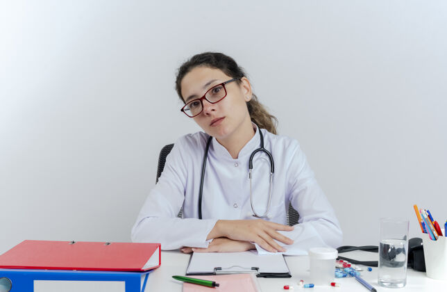 穿年轻的女医生穿着医用长袍 戴着听诊器和眼镜 坐在办公桌旁 手里拿着医疗工具 手放在办公桌上 看上去很孤立年轻长袍人