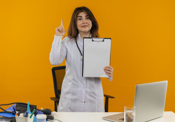 听诊器印象深刻的中年女医生穿着医用长袍和听诊器站在办公桌后面 拿着医疗工具和笔记本电脑拿着剪贴板 抬起手指隔离医疗笔记本电脑穿着