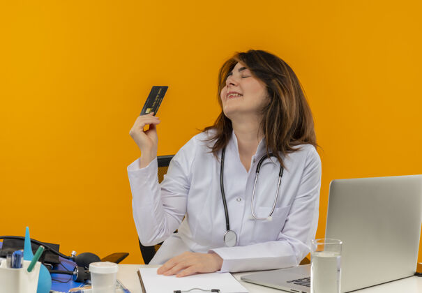信用卡高兴的中年女医生穿着医用长袍 听诊器坐在办公桌旁 带着医疗工具剪贴板和笔记本电脑 抱着信用卡 闭着眼睛 与世隔绝橙色中年听诊器