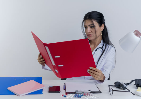 长袍皱眉的年轻女医生穿着医用长袍和听诊器坐在办公桌旁 手持医疗工具 看着文件夹人年轻人成人