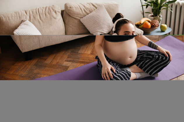 房间照片中 身穿上衣的晒黑孕妇坐在紫色瑜伽地毯上 戴着耳机听音乐哑铃快乐感觉
