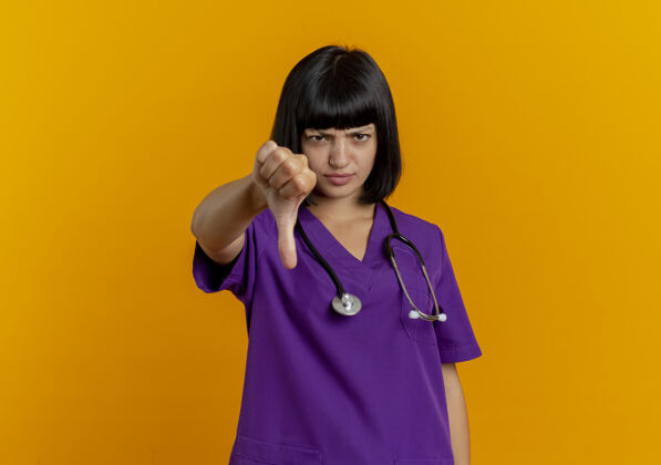 太空年轻的黑发女医生穿着制服 听诊器大拇指朝下 橙色背景 留有复印空间不愉快女性制服