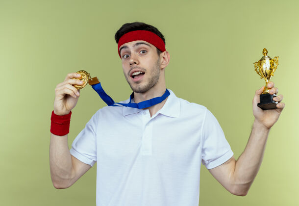 奖牌戴着头巾 脖子上戴着金牌 手持奖杯的年轻运动型男子站在绿色的背景上 快乐而兴奋抱着头带兴奋