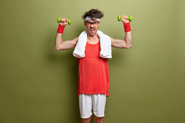 年轻健康的生活方式和体育锻炼意志坚定的男人举起哑铃锻炼肌肉 全力以赴取得积极的结果 穿着运动服 脖子上围着毛巾 在绿色隔离毛巾健身房决心
