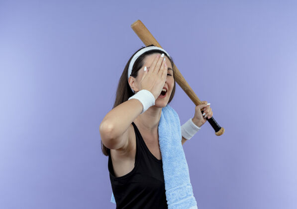 微笑戴着头巾 肩上扛着毛巾 手持棒球棒 一只手微笑着站在蓝色背景上的年轻健身女士毛巾眼睛手